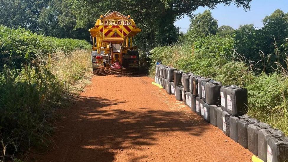 A council grit lorry spreading what appears to be sand on a narrow country lane. There is a pile of oil drums piled up on the right hand side of the road. There are trees and hedgerows either side.