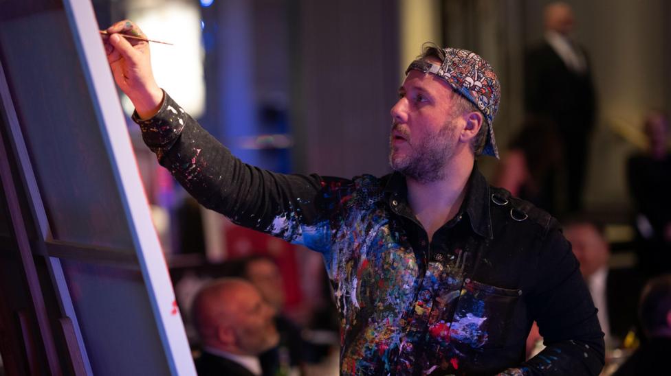 A man with a backwards cap painting, his shirt is covered in paint and he is looking at a canvas holding a paintbrush in his right hand.