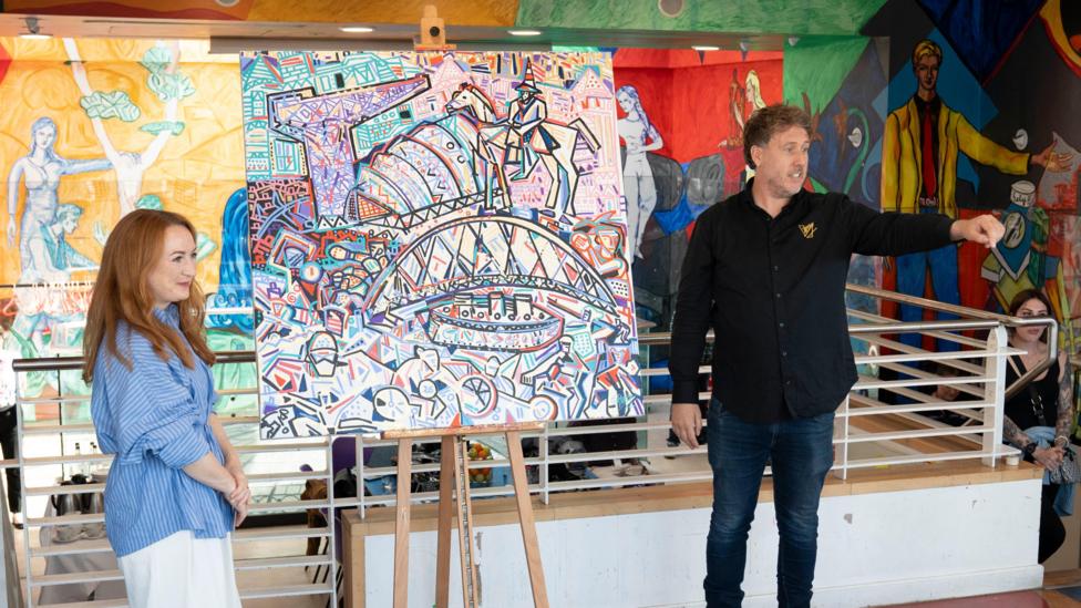 A painting in the middle of the photograph, with a ginger woman on the left hand side in a blue shirt and white trousers and her arms crossed. On the right hand side of the painting is a man in a black shirt and dark blue jeans looking over to the right side of him and pointing.