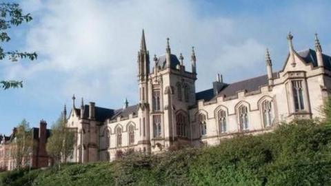 The university campus in Londonderry. A number of bushes and grassy areas can be seen in front of a Gothic-looking building with several large windows.