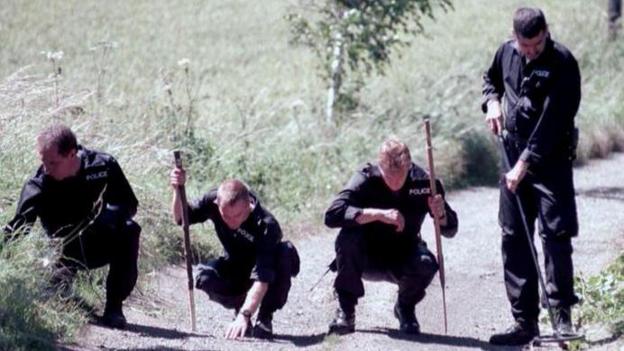 Four police officers in a line, looking for evidence on a country lane. They are dressed in black police overalls and two are carrying sticks. Three are crouched down looking at the ground, while one is standing with what appears to be a metal detector.