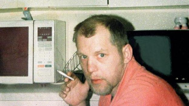 Michael Stone, holding a cigarette in his right hand and looking towards the camera. He has heavy stubble and is wearing a peach-coloured top. A microwave oven can be seen behind him.