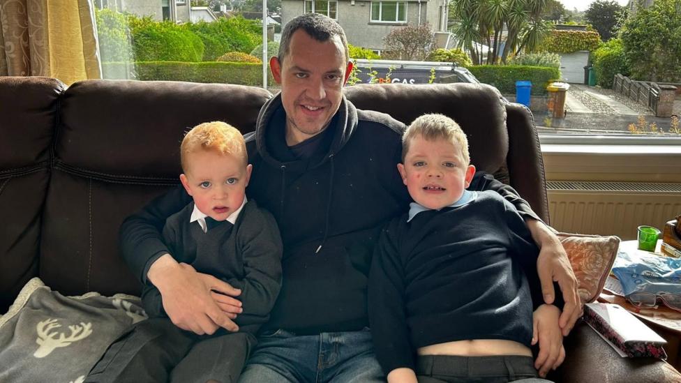 A person in a black hoodie sits on a brown leather sofa between two young children wearing school uniforms. The three are positioned in front of a large picture window overlooking a residential street with houses, hedges, and trees. 
