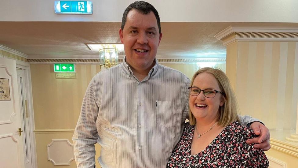 Two people stand together in a hotel or function venue corridor with cream striped walls and a red patterned carpet. One person wears a light striped button-down shirt and jeans, while the other wears a dark floral dress and glasses.