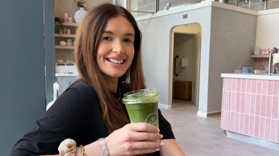 Jess smiles, holding an iced matcha latte in a cafe. She has long brown hair and wears a black top