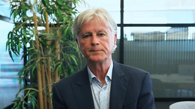 James, a man in a dark suit jacket and light shirt sits in an office space with tall buildings visible through the windows and a plant behind him. He has white hair and looks at the camera.