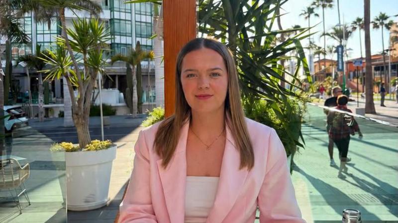 Mia is pictured in a sunny climate. She's wearing a light pink blazer with a white top underneath and a dainty necklace. She has light brown hair.