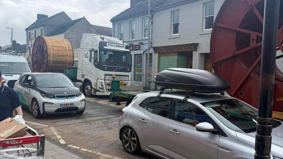 The main road through Sanquhar clogged up with traffic, we can see cars in the foreground and a lorry with a large, round, wooden cog on the back in the background. The vehicles appear to be at a standstill.