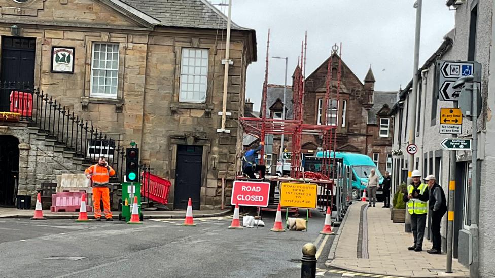 A picture of road works in Sanquhar with scaffolding out on the roadway next to the historic Tolbooth Museum on the left. There is a road closed sign and traffic lights and a man in orange overalls on the phone and another in a high-visibility jacket and hard hat talking to a member of the public