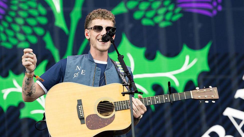 Nathan Evans on a stage in front of a microphone and playing the guitar. He is wearing a blue t-shirt, denim gilet and dark sunglasses. A thistle is projected on a screen in the background.