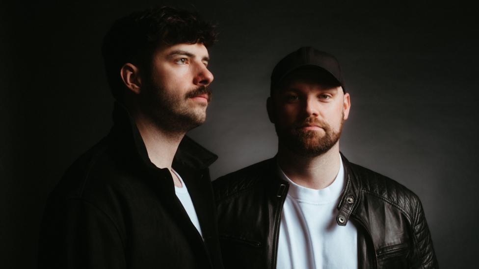 Two men with dark hair, beards and moustaches wearing a white t-shirts and black jackets. One is facing forward and looking at the camera while the other is looking to the right away from the camera.