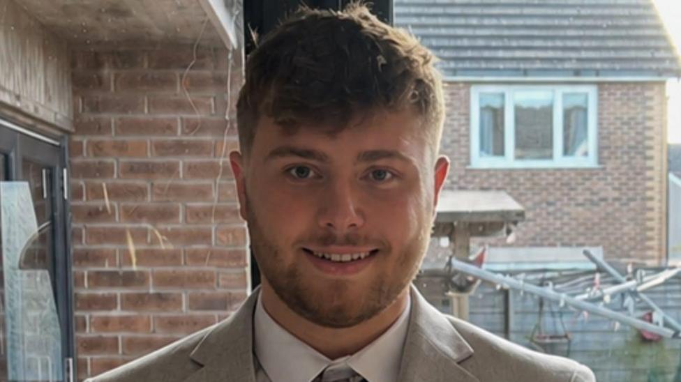 Ryan Davies yn gwisgo siwt brown ac yn gwenu at y camera