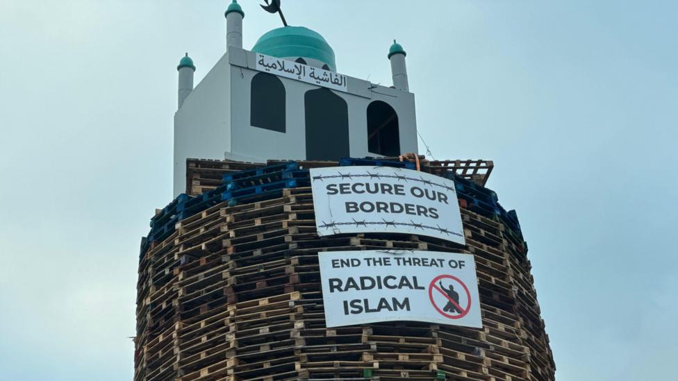 An image of the top of the bonfire, consisting of wood pallets, with a structure resembling a mosque on top. Two placards are below. One reads