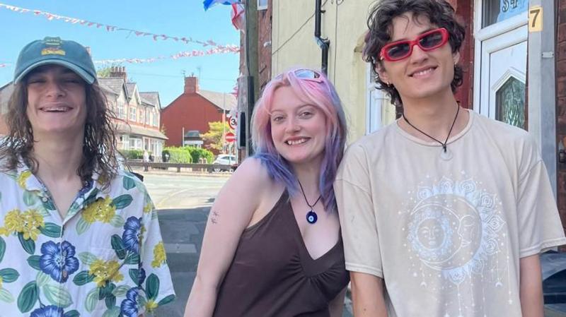 Three teengers, two men and one woman, smile at the camera on a street with flags and bunting in the background
