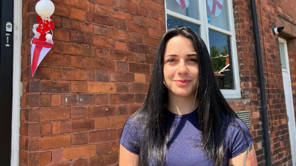 A young woman will long, straight black hair and wearing a purple t-shirt smiles at the camera outside a terraced house decorated with England flags