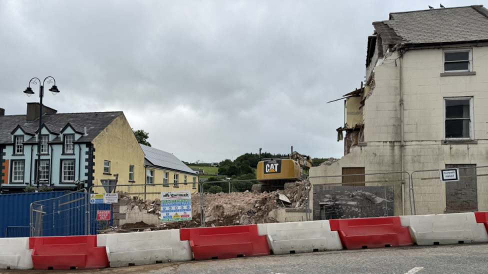 A large digger is pictured over the rubble of a building. A large gap is visible between two exterior buildings and a number of cordons are in place