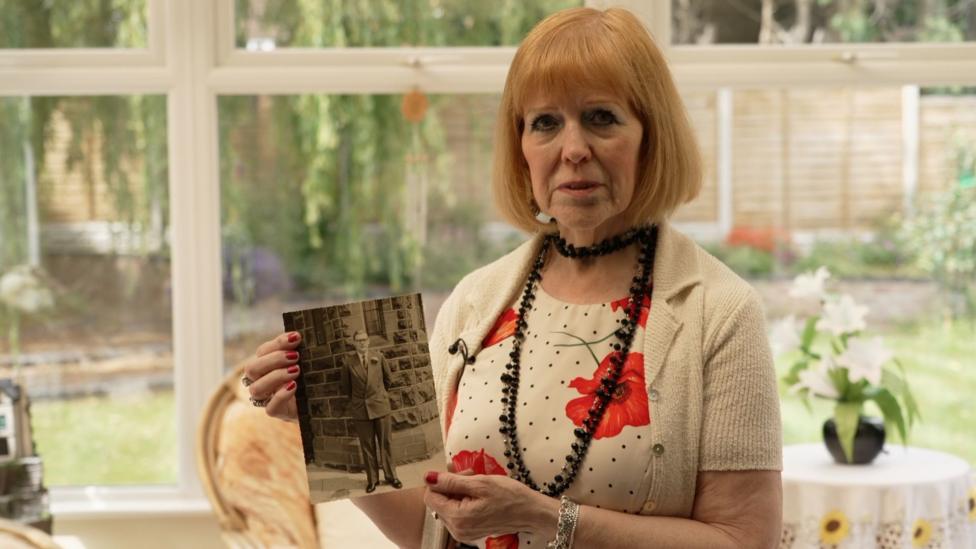 A woman with auburn hair, wearing a cream cardigan, a flower-patterned top and a black beaded necklace, stands in a bright conservatory holding up a small black-and-white photograph of a man in a suit. Her expression is serious.