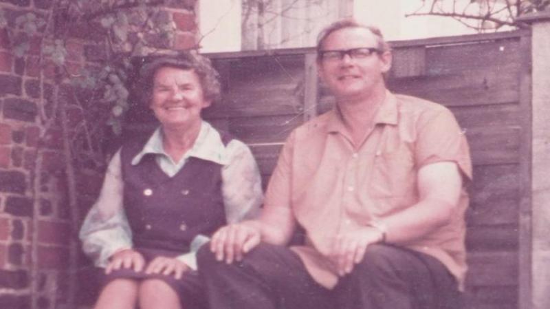 A faded colour photograph of Anthony Littler and an older woman sitting side by side on a bench in front of a brick wall and wooden fence. He wears glasses and a short-sleeved shirt; she wears a dark pinafore over a white blouse. Both are smiling.