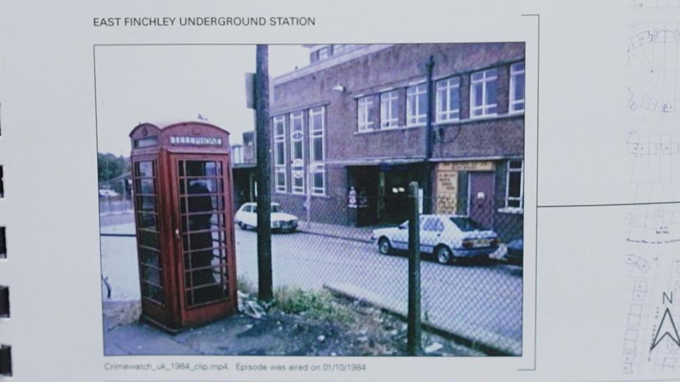 A colour 1984 photograph of a traditional red telephone box standing on a street corner, with parked cars and low buildings behind it.