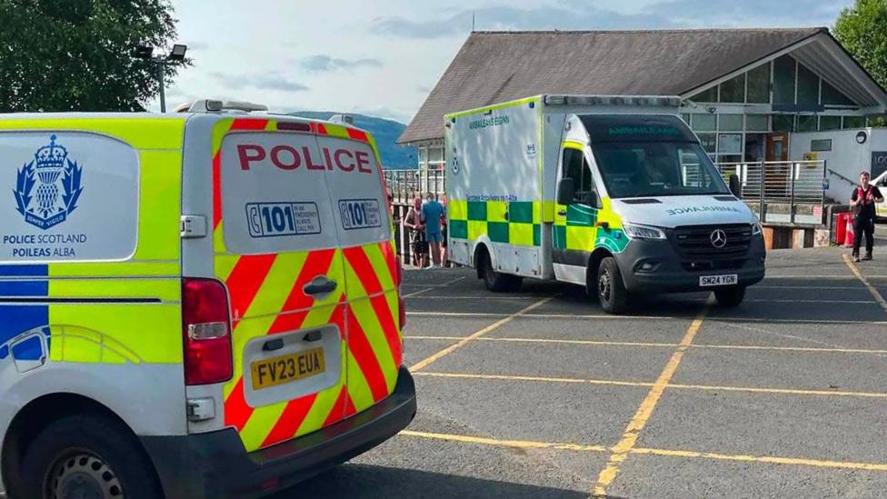 A police van and an ambulance in a car park outside a building with railings. A hill is visible in the background.