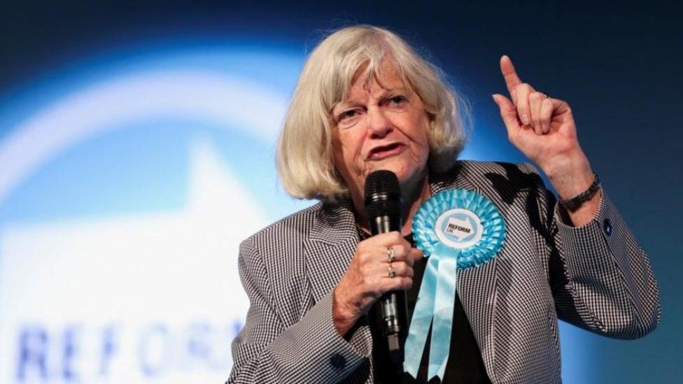 Ann Widdecombe speaks on the stage during the Reform UK party's rally at the NEC in Birmingham, Britain, June 30, 2024. She is wearing a black and white houndstooth jacket with a large turquoise Reform UK rosette. She is holding a microphone and gesticulating, with a serious expression. Her hair is worn in a blonde bob with a fringe, and she is also wearing some rings on ger fingers and a watch.