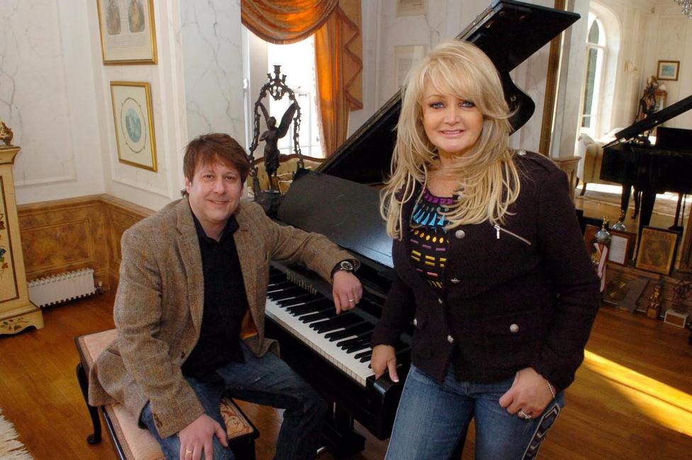 A man with brown hair, wearing blue jeans, a black t shirt and a brown jacket. He is sat at a piano. Stood up beside him is a glamorous blonde woman wearing blue jeans and a black top and jacket. They are both looking at the camera and smiling.
