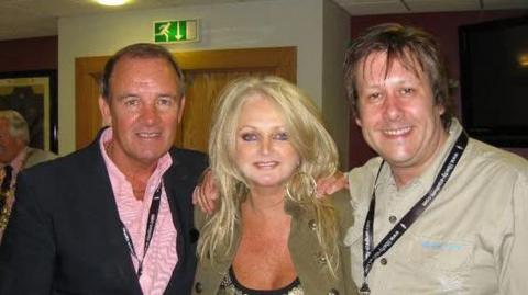 A glamorous blonde woman with two men on either side of her. They are all smiling and looking at the camera.