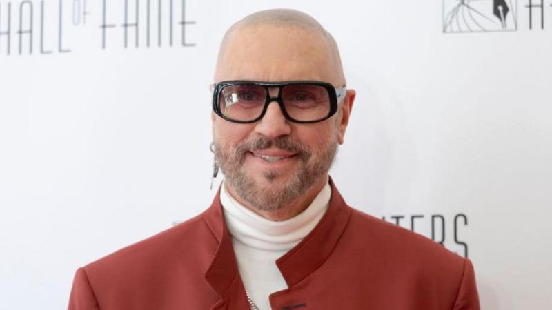 A bald man with grey facial hair wearing one earring and sunglasses, as well as a white polo kneck jumper and red jacket.