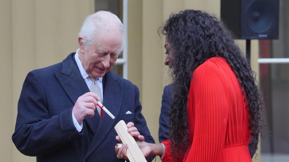 King Charles wearing a dark suit and grey tie places a message into a wooden baton. A woman with dark curly hair and a red dress holds the baton. 