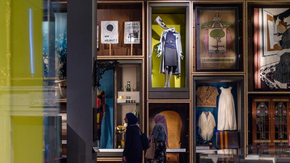 A musem display which shows mannequins of clothes on display in the windows, and artwork. The display shows windows with items in the windows. There are two women walking through the display.