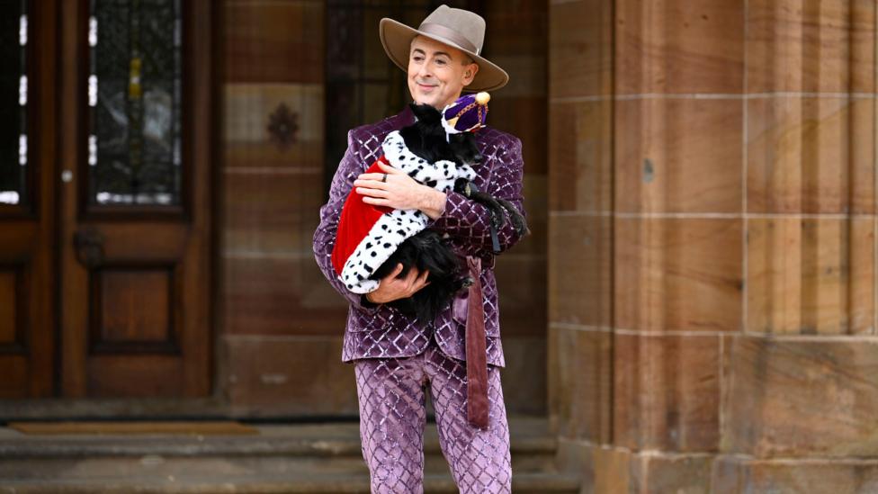 Alan Cumming, a man, in a purple diamond quilted suit, a grey suede cowboy hot, stood central to the photo infront of a building and he is smiling looking to the left side of the photo. He is cradling a black dog in his arms, the dog is wearing a crown and has a royal cape on. 