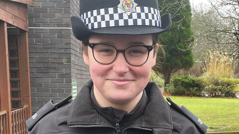 Jess Turnbull, wearing police uniform and smiling. She is standing in front of a dark grey brick building with a wooden entrance to one side. Behind her to the other side is a patch of grass, a tall, thin bush-like tree and some other trees, like you might find in the gardens of an office building. Jess is wearing a rounded police hat and black rimmed glasses and has her dark hair tied back.