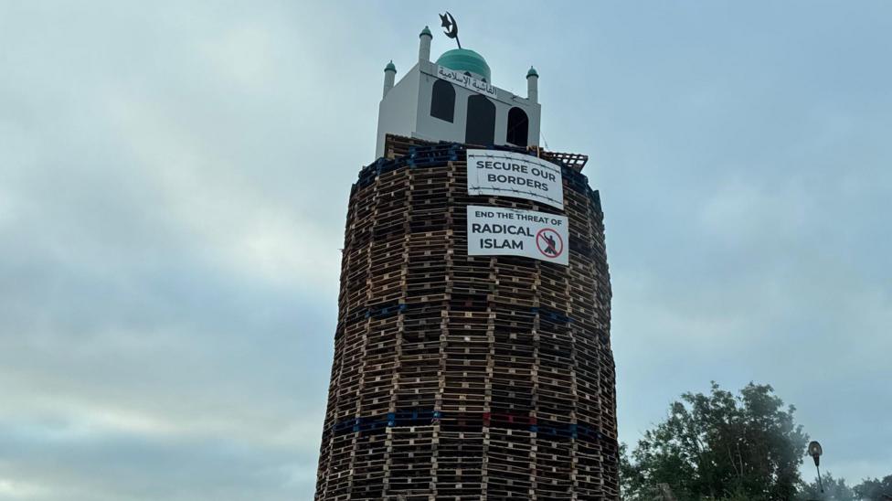 An effigy of a mosque is on top of a bonfire built out of wooden pallets. Signs say secure our vorder and end the threat of radical Islam. The sky is cloudy.