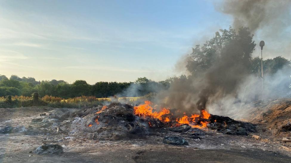 The burnt out remains of a bonfire. It's still smouldering with fire close to the ground. Black ash is lying on the ground. Smoke is rising out of the fire. Trees are in the background.