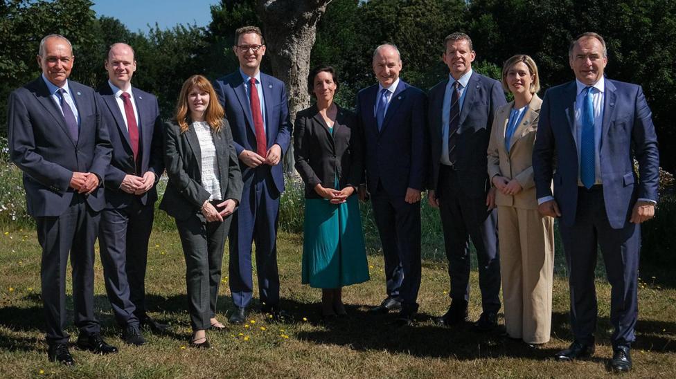 Six white middle-aged men in dark blue or navy suits and three smartly dressed women standing together in a garden. They are all looking at the camera smiling. There are trees in the background and blue skies.