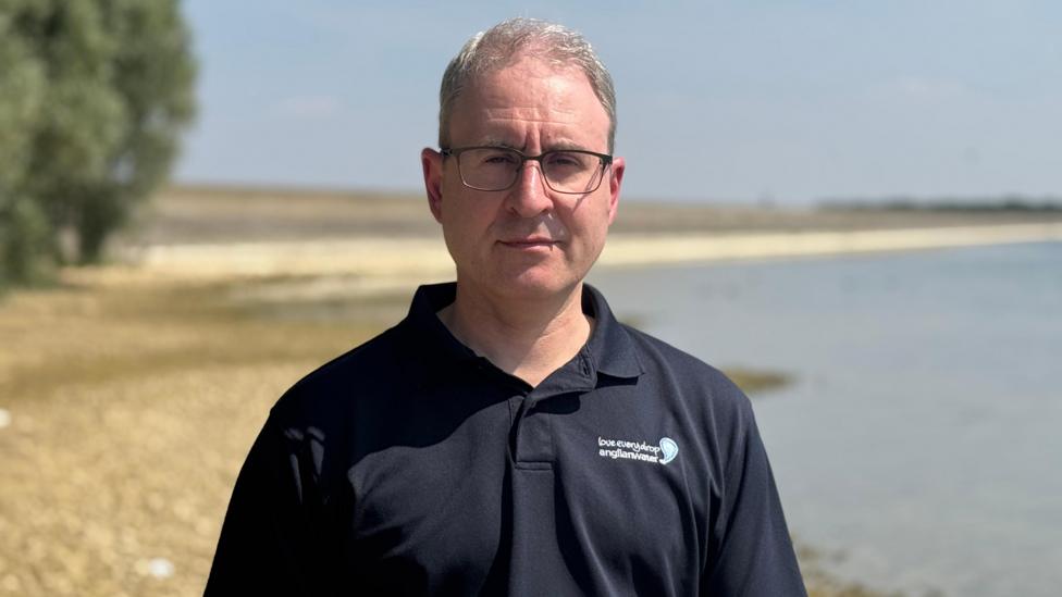 Geoff looks at the camera with a fairly serious expression. He wears a navy blue collared casual shirt with the Anglian water logo that says