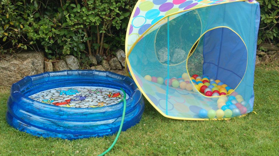 A blue plastic paddling pool in the garden, with a green hose running into it. It is next to a mini tent full of plastic lightweight balls.