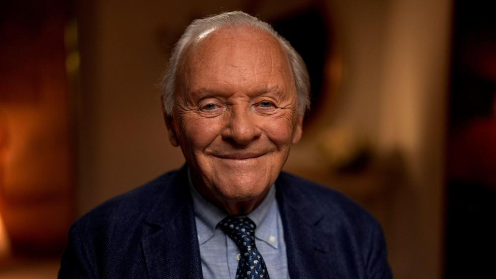 Sir Anthony Hopkins looking to camera and smiling; dressed in navy blue suit, light blue shirt, and dark blue and white tie