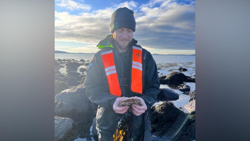 Dr Nick Baker Horne is wearing a beanie hat and wax coat and orange scarf. He is standing in a coastal area holding a crab