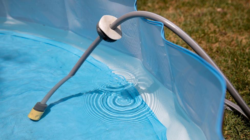 A hosepipe is laying in a blue paddling pool which is sitting on grass. Water has filled up the pool half way.