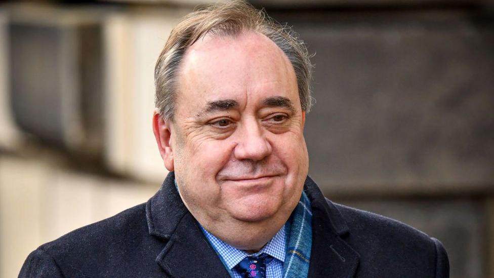 A smiling man with receding hair is wearing a dark coat, tartan scart, blue shirt and tie