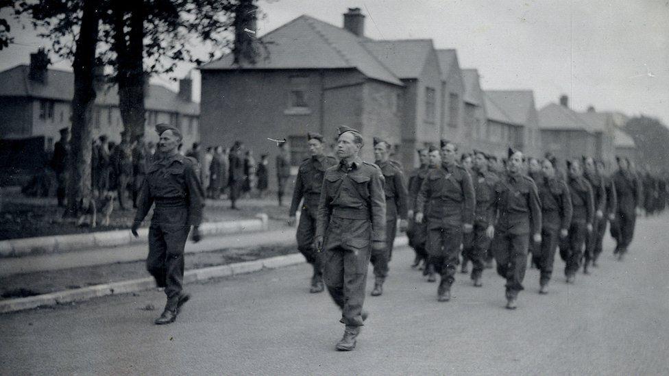 A black and white picture of men marching past houses