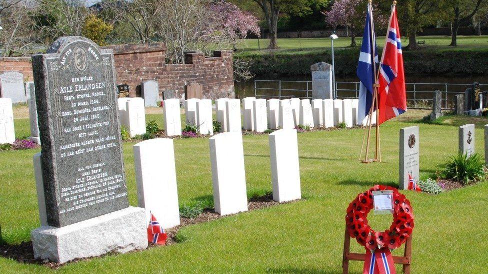 A cemetery with Scotland and Norway flags intertwined and gravestones across the grass and a wreath of poppies with Norwegian colours attached