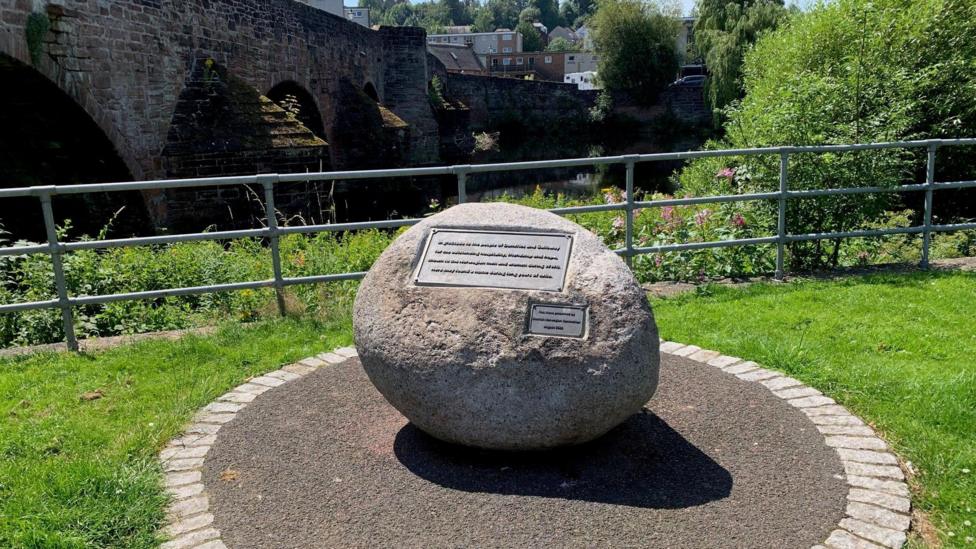 A large stone with a metal panel on it sits on tarmac beside a grassy area and a gate next to a bridge over the River Nith