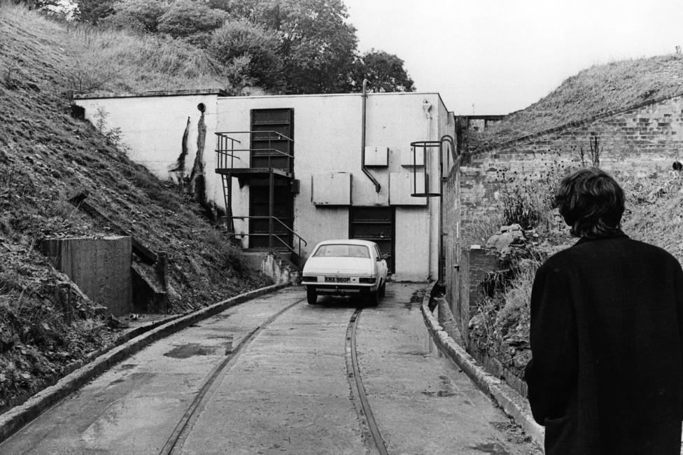 Black-and-white photograph of a concrete service building set into a hillside, resembling a bunker or underground facility entrance. A narrow roadway with rail tracks embedded in the surface leads to the building, where a small car is parked outside. In the foreground, a person wearing a dark coat is seen from behind looking toward the structure. Steep grassy banks and retaining walls flank the entrance, creating a secluded, industrial setting.
