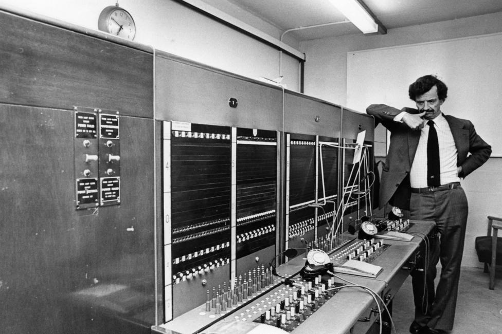 A black-and-white photograph of a telecommunications room. A man with dark hair and a moustache stands at the far end of a large telephone switchboard that stretches across most of the image. The man is wearing a black suit. The switchboard is covered with rows of connection points, switches and patch cords, while several telephones sit on the work surface below.