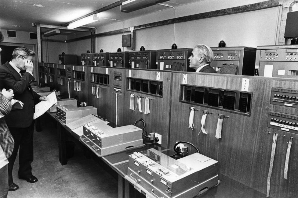 Black-and-white photograph of a room filled with telecommunications or computer equipment. Large cabinets line the walls, with control panels, switches and labelled tape or paper strips hanging from them. Several people stand among the equipment, examining documents and the machinery. Long worktables in the foreground hold electronic devices and monitoring equipment, illustrating a mid-20th-century technical operations centre.