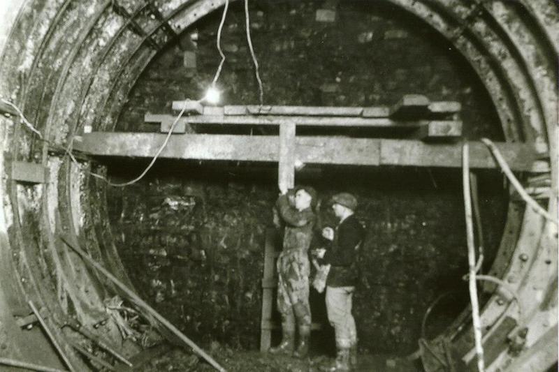 A black and white photo of two men building a tunnel, a brick wall is visible at the end and a metal tube is. There is tubes and wires hanging down.