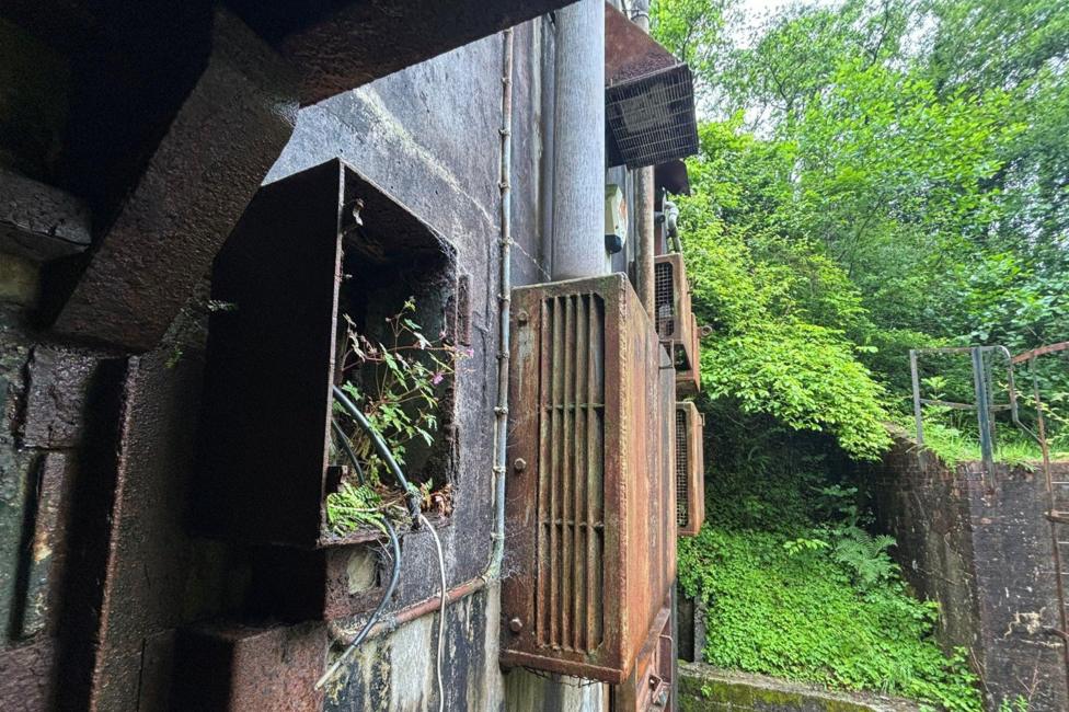 A rusty exterior of the bunker with wires protruding. The bunker looks overgrown.