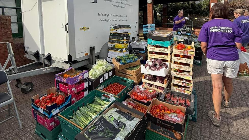A large selection of boxes of fresh food, all piled up, next to three people who are wearing purple T-shirts with the words Larder Volunteer Food Hero in white writing on the back. There is a white trailer unit to the left.